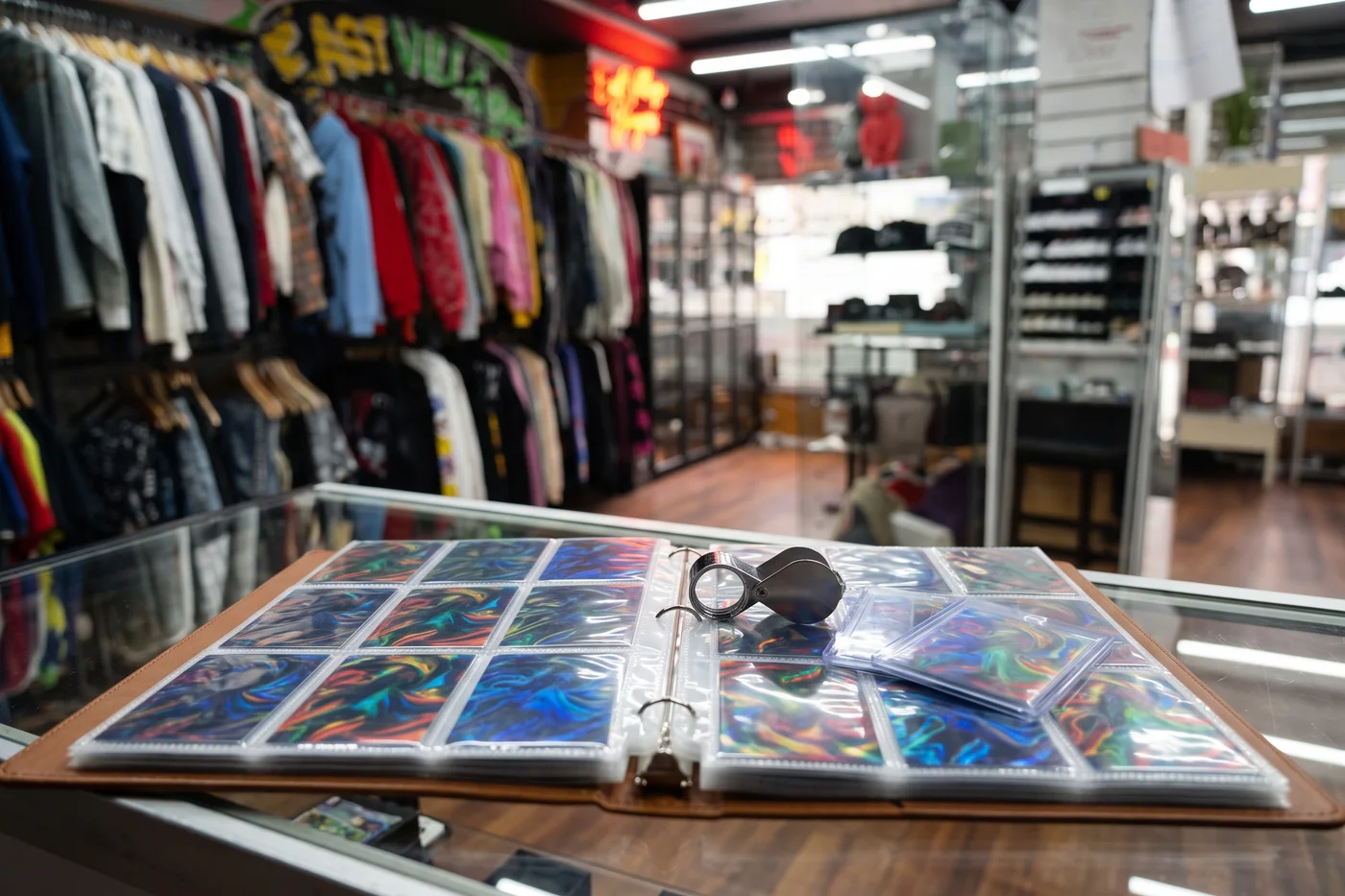 An open trading card binder with holographic cards, a jeweler's loupe and cards in toploaders on the counter at East Village Buyers